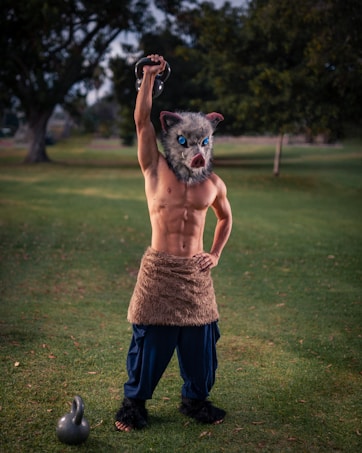 A muscular person stands on grass in a park, wearing a wolf mask and a furry waist cloth, with a kettlebell in one hand raised above their head and another kettlebell on the ground nearby.