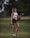 A fit woman stands on a grassy field holding two dumbbells, one in each hand. She is wearing a white sports bra, black shorts, and sneakers. The background includes soft-focused trees and foliage, contributing to a serene, outdoor setting.