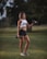 A fit woman stands on a grassy field holding two dumbbells, one in each hand. She is wearing a white sports bra, black shorts, and sneakers. The background includes soft-focused trees and foliage, contributing to a serene, outdoor setting.