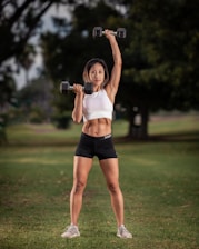 A fit individual is performing an exercise with dumbbells outdoors on a grassy field. The person is wearing a white crop top and black shorts, holding one dumbbell in each hand.