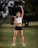 A fit individual is performing an exercise with dumbbells outdoors on a grassy field. The person is wearing a white crop top and black shorts, holding one dumbbell in each hand.