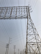 A series of tall metal power lines and pylons create a complex network against a clear sky. The structures feature intricate, geometric patterns with many intersecting lines.