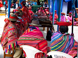 People are gathered in a market setting, wearing traditional, colorful woven garments with intricate patterns. They are seated on white plastic chairs, and their hats are adorned with red bands. In the background, there are several booths or tables covered in textiles and ornaments. Some individuals are standing or walking around.