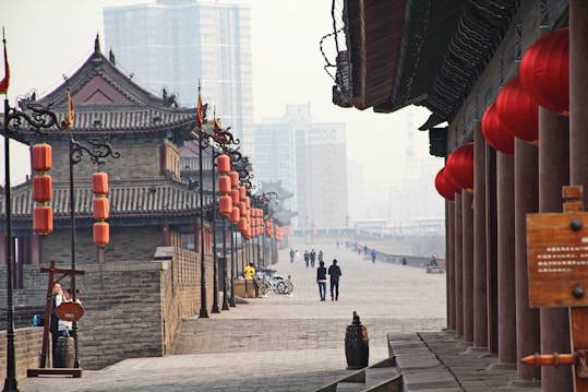A historical city wall with traditional architecture featuring stone walls and ornate wooden structures lined with red lanterns. Modern skyscrapers rise in the background, creating a contrast between old and new. People are walking along the path, and several bicycles are parked nearby.