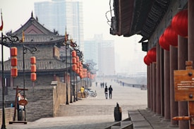A historical city wall with traditional architecture featuring stone walls and ornate wooden structures lined with red lanterns. Modern skyscrapers rise in the background, creating a contrast between old and new. People are walking along the path, and several bicycles are parked nearby.