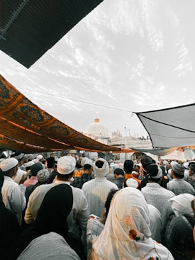A large gathering of people dressed in traditional attire, many of whom are wearing head coverings. They appear to be facing a structure with a golden dome, possibly a religious or cultural event. Above, there are decorative cloths providing shade, with a partially cloudy sky visible.
