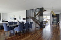 A modern, open-concept dining and living area featuring a large wooden dining table surrounded by blue cushioned chairs. The room has wooden flooring and a contemporary staircase with vertical slender railing slats leading to an upper level. Artistic wall decorations and elegant lighting fixtures add to the sophisticated atmosphere. Large windows allow natural light to fill the space, enhancing its airy and spacious feel.