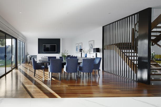 A modern dining area with a long table surrounded by blue upholstered chairs sits on polished wooden flooring. The space is well-lit with natural light streaming through large glass doors on the left. A flat-screen TV is mounted on a black accent wall, and a contemporary staircase with wooden steps features a sleek black railing. Decorative elements like vases with flowers are placed throughout the room.