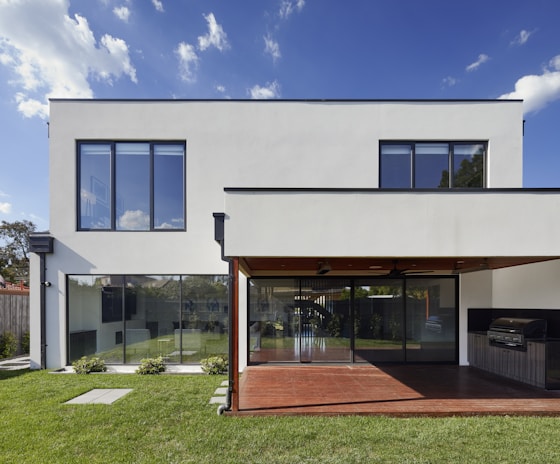 A modern two-story house with large rectangular windows and a minimalist design. The exterior is white with a clean, geometric structure, and there is a glass sliding door leading to a covered patio area. The patio features a built-in stainless steel grill, and the yard is well-manicured with green grass and small shrubs.