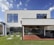 A modern two-story house with large rectangular windows and a minimalist design. The exterior is white with a clean, geometric structure, and there is a glass sliding door leading to a covered patio area. The patio features a built-in stainless steel grill, and the yard is well-manicured with green grass and small shrubs.