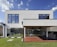 A modern two-story house with large rectangular windows and a minimalist design. The exterior is white with a clean, geometric structure, and there is a glass sliding door leading to a covered patio area. The patio features a built-in stainless steel grill, and the yard is well-manicured with green grass and small shrubs.