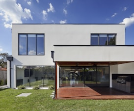 A modern two-story house with large rectangular windows and a minimalist design. The exterior is white with a clean, geometric structure, and there is a glass sliding door leading to a covered patio area. The patio features a built-in stainless steel grill, and the yard is well-manicured with green grass and small shrubs.