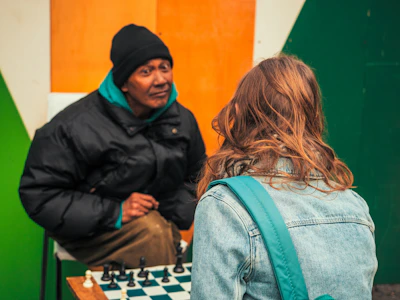 Two people are outdoors, engaged in a game of chess. One person is wearing a black jacket and beanie, while the other is dressed in a denim jacket with long hair. A colorful backdrop with green and orange sections frames the scene, and a chessboard with pieces is placed on a small wooden table between them.