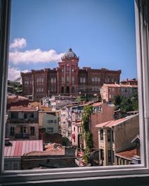 A view through an open window reveals a historic red-brick building with a dome, surrounded by a series of smaller buildings with varying architectural styles. The sky is clear and blue, indicating a sunny day. Greenery is visible on some rooftops and a Turkish flag hangs from one of the buildings.