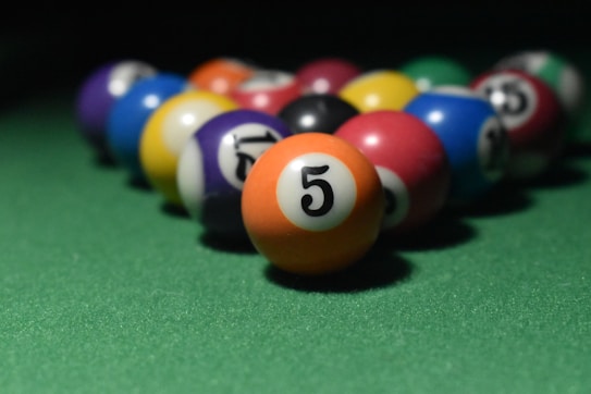 A collection of colorful billiard balls arranged on a green pool table, with the number 5 ball prominently in the foreground. The lighting creates a soft shadow, emphasizing the vibrant colors of the balls.
