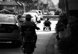 A person in a wheelchair is moving along a city street amidst traffic, including a taxi and motorbike. The scene is in black and white, capturing an urban environment with vehicles and buildings in the background. The image is rich in light contrast, giving it a dramatic look.