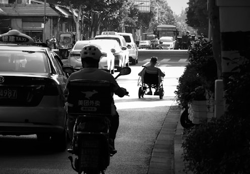 A person in a wheelchair is moving along a city street amidst traffic, including a taxi and motorbike. The scene is in black and white, capturing an urban environment with vehicles and buildings in the background. The image is rich in light contrast, giving it a dramatic look.
