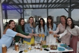 A group of seven women standing together at an outdoor cafe or gathering spot, smiling and toasting with glasses of orange juice. They are dressed casually with a variety of clothing styles. The setting includes a table with plates, food items, and glasses in the foreground. In the background, large windows reflect the cityscape scenery.