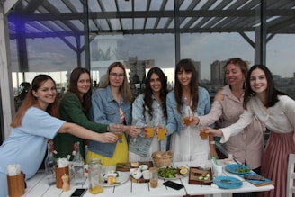A group of seven women standing together at an outdoor cafe or gathering spot, smiling and toasting with glasses of orange juice. They are dressed casually with a variety of clothing styles. The setting includes a table with plates, food items, and glasses in the foreground. In the background, large windows reflect the cityscape scenery.