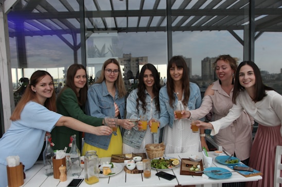 A group of seven women standing together at an outdoor cafe or gathering spot, smiling and toasting with glasses of orange juice. They are dressed casually with a variety of clothing styles. The setting includes a table with plates, food items, and glasses in the foreground. In the background, large windows reflect the cityscape scenery.