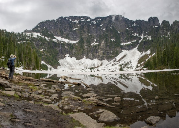 A tranquil mountain landscape with snow patches scattered across steep rocky cliffs reflected in a calm lake. A lone hiker stands on the rocky shore, gazing towards the mountains while wearing a backpack and wide-brimmed hat. Evergreen trees line the lakeside, blending into the natural scenery under a cloudy sky.