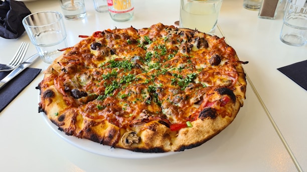 A freshly baked pizza topped with cheese, mushrooms, olives, and green herbs. It is presented on a white plate on a light-colored table. Surrounding the pizza are various dining items, including glasses, cutlery, and napkins.