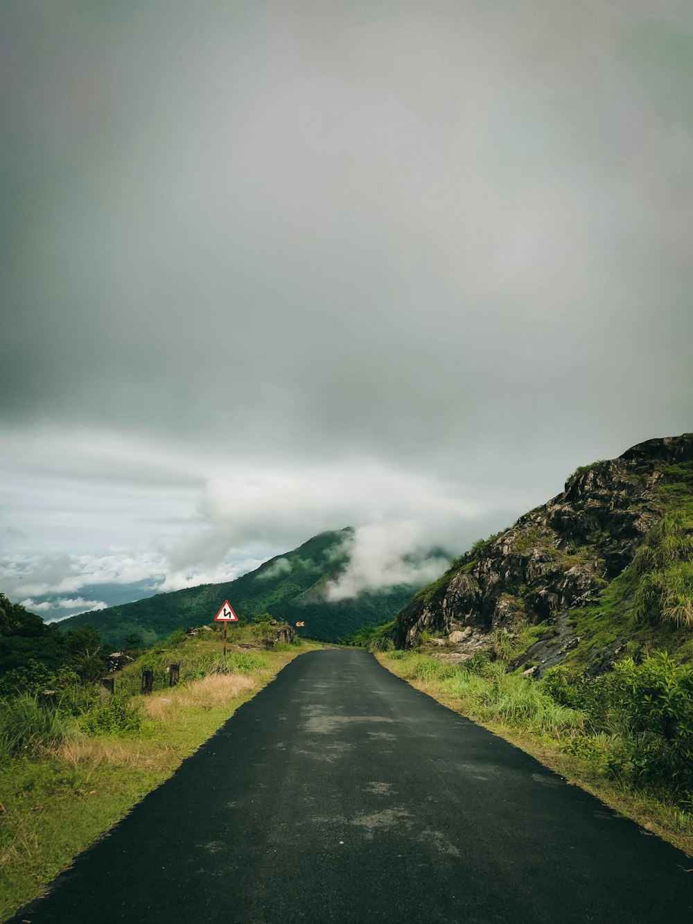 Route en béton gris entre un champ d’herbe verte près de la montagne sous des nuages gris pendant la journée