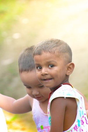 Two young children are captured in a moment of interaction. The child in the foreground is smiling gently, while the other child is partially visible and looking down. They are wearing light-colored sleeveless tops. The background is softly blurred with natural outdoor lighting, adding a warm ambiance.