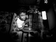 A young child is sitting on a bed using a desktop computer. The child is smiling and wearing a cap, with one hand on the keyboard and the other holding a computer mouse. The bedspread features a floral pattern, and the room is dimly lit, adding a dramatic contrast to the scene.