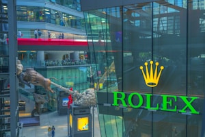 A vibrant shopping mall scene with a large Rolex logo prominently displayed on a glass window. The interior architecture features multiple levels with balconies and escalators. A unique sculpture of a bull is part of the decor, and people are visible walking and interacting throughout the space.