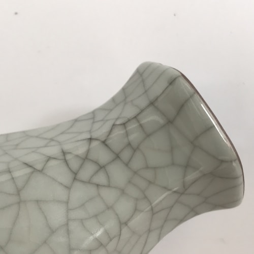 A close-up of a ceramic object featuring a distinctive craquelure pattern, with a glossy surface reflecting light. The object has a curved shape and appears to be a part of a vase or a ceramic piece.