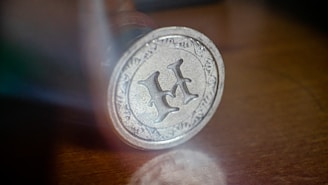 A detailed close-up of a metal seal with the letter 'H' prominently displayed in the center. The seal is circular with an ornate border featuring intricate floral patterns. The surface of the metal has a slightly tarnished appearance, suggesting age or frequent use. The surrounding area is blurred, focusing attention on the seal itself, which rests on a wooden surface.
