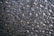The image depicts a close-up view of a textured surface, which appears to be a frosted or patterned glass. The surface has an irregular, geometric pattern with raised areas that create a sense of depth and complexity. The colors are predominantly dark, with shades of black and grey, interspersed with occasional warm highlights that might be reflections.