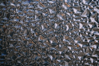 The image depicts a close-up view of a textured surface, which appears to be a frosted or patterned glass. The surface has an irregular, geometric pattern with raised areas that create a sense of depth and complexity. The colors are predominantly dark, with shades of black and grey, interspersed with occasional warm highlights that might be reflections.