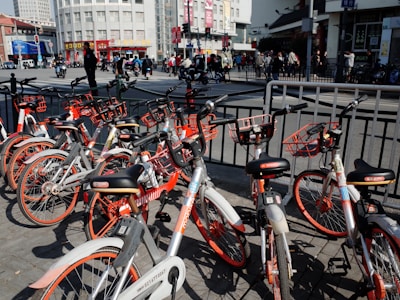 A collection of rental bicycles with orange accents is parked in a row on a city street. The background shows a busy urban environment with people walking and vehicles on the move. Tall buildings and signage are visible, and there are railings separating the bicycles from the road.