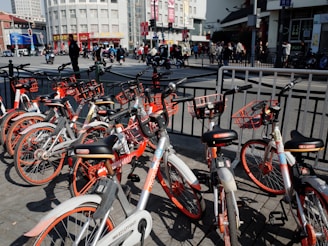 A collection of rental bicycles with orange accents is parked in a row on a city street. The background shows a busy urban environment with people walking and vehicles on the move. Tall buildings and signage are visible, and there are railings separating the bicycles from the road.