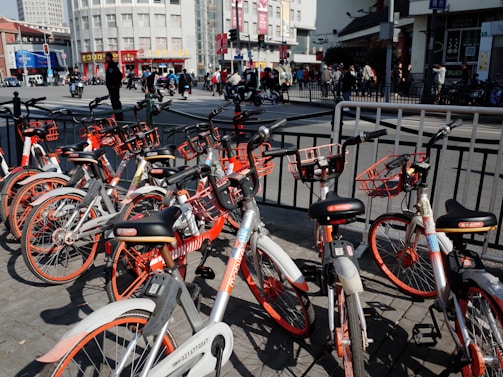 A collection of rental bicycles with orange accents is parked in a row on a city street. The background shows a busy urban environment with people walking and vehicles on the move. Tall buildings and signage are visible, and there are railings separating the bicycles from the road.
