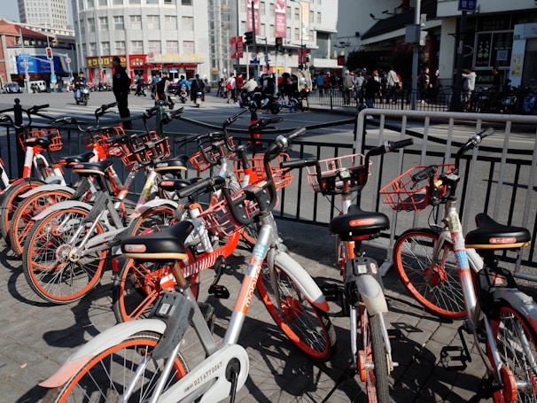 A collection of rental bicycles with orange accents is parked in a row on a city street. The background shows a busy urban environment with people walking and vehicles on the move. Tall buildings and signage are visible, and there are railings separating the bicycles from the road.