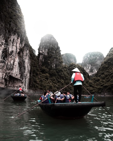 A group of people in life jackets are seated in a boat on calm water, surrounded by towering limestone cliffs covered in lush greenery. The boat is being maneuvered by a person standing with an oar, wearing a conical hat and a red vest.
