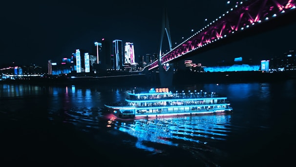 A brightly lit boat cruises on a river at night, passing beneath a bridge adorned with pink and purple lights. The background features a city skyline with tall buildings illuminated by vibrant blue and white lights, reflecting on the water’s surface.