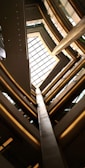An architectural view looking up through multiple floors of a building, featuring a large glass ceiling that lets in natural light. The structure is composed of geometric lines and angles, with metal and glass as prominent materials.