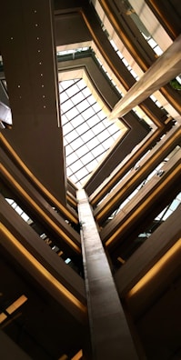 An architectural view looking up through multiple floors of a building, featuring a large glass ceiling that lets in natural light. The structure is composed of geometric lines and angles, with metal and glass as prominent materials.