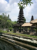 A traditional Balinese temple complex with tiered pagodas surrounded by lush green foliage and stone structures. The sky is partly cloudy, adding a serene atmosphere to the scene. The intricate architecture and thatched roofs exhibit cultural significance, with trees and plants providing a natural backdrop.