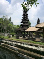 A traditional Balinese temple complex with tiered pagodas surrounded by lush green foliage and stone structures. The sky is partly cloudy, adding a serene atmosphere to the scene. The intricate architecture and thatched roofs exhibit cultural significance, with trees and plants providing a natural backdrop.