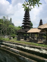 A traditional Balinese temple complex with tiered pagodas surrounded by lush green foliage and stone structures. The sky is partly cloudy, adding a serene atmosphere to the scene. The intricate architecture and thatched roofs exhibit cultural significance, with trees and plants providing a natural backdrop.