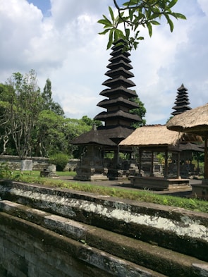 A traditional Balinese temple complex with tiered pagodas surrounded by lush green foliage and stone structures. The sky is partly cloudy, adding a serene atmosphere to the scene. The intricate architecture and thatched roofs exhibit cultural significance, with trees and plants providing a natural backdrop.