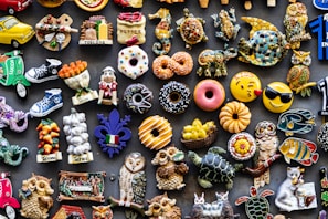 A colorful assortment of decorative fridge magnets depicting various objects such as donuts, animals, owls, fruits, and Italian-themed items. The magnets are arranged closely together on a dark background, showcasing a wide variety of shapes and designs, including smiley faces and motorcycles.