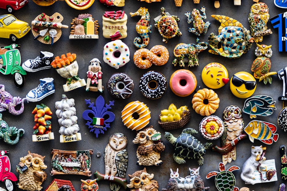 A colorful assortment of decorative fridge magnets depicting various objects such as donuts, animals, owls, fruits, and Italian-themed items. The magnets are arranged closely together on a dark background, showcasing a wide variety of shapes and designs, including smiley faces and motorcycles.