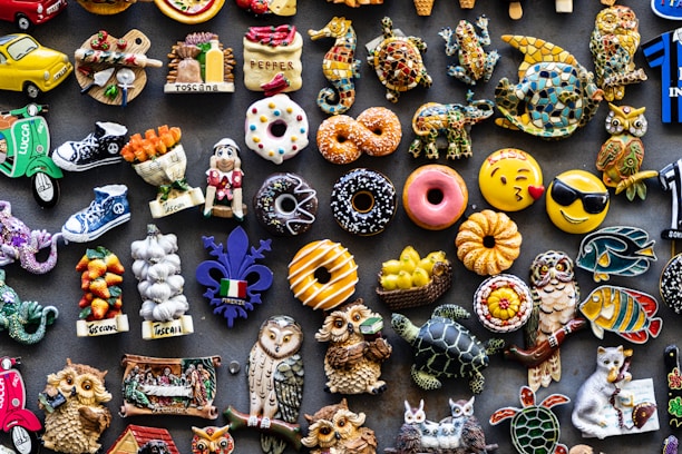 A colorful assortment of decorative fridge magnets depicting various objects such as donuts, animals, owls, fruits, and Italian-themed items. The magnets are arranged closely together on a dark background, showcasing a wide variety of shapes and designs, including smiley faces and motorcycles.