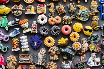 A colorful assortment of decorative fridge magnets depicting various objects such as donuts, animals, owls, fruits, and Italian-themed items. The magnets are arranged closely together on a dark background, showcasing a wide variety of shapes and designs, including smiley faces and motorcycles.